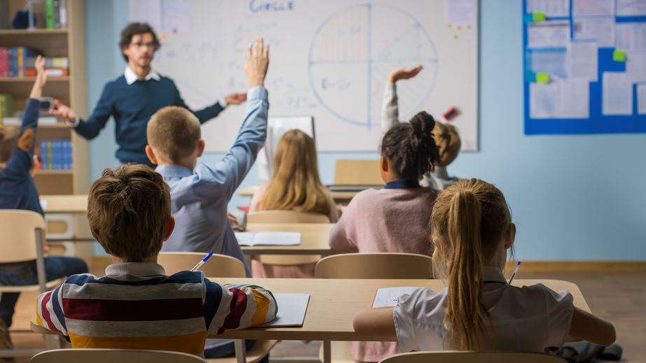 Caring Teacher Explains Lesson to a Classroom Full of Diverse Children. In School with Group of Smart Multiethnic Kids Learning Science, Whole Classroom Raising Hands Knowing Answer.